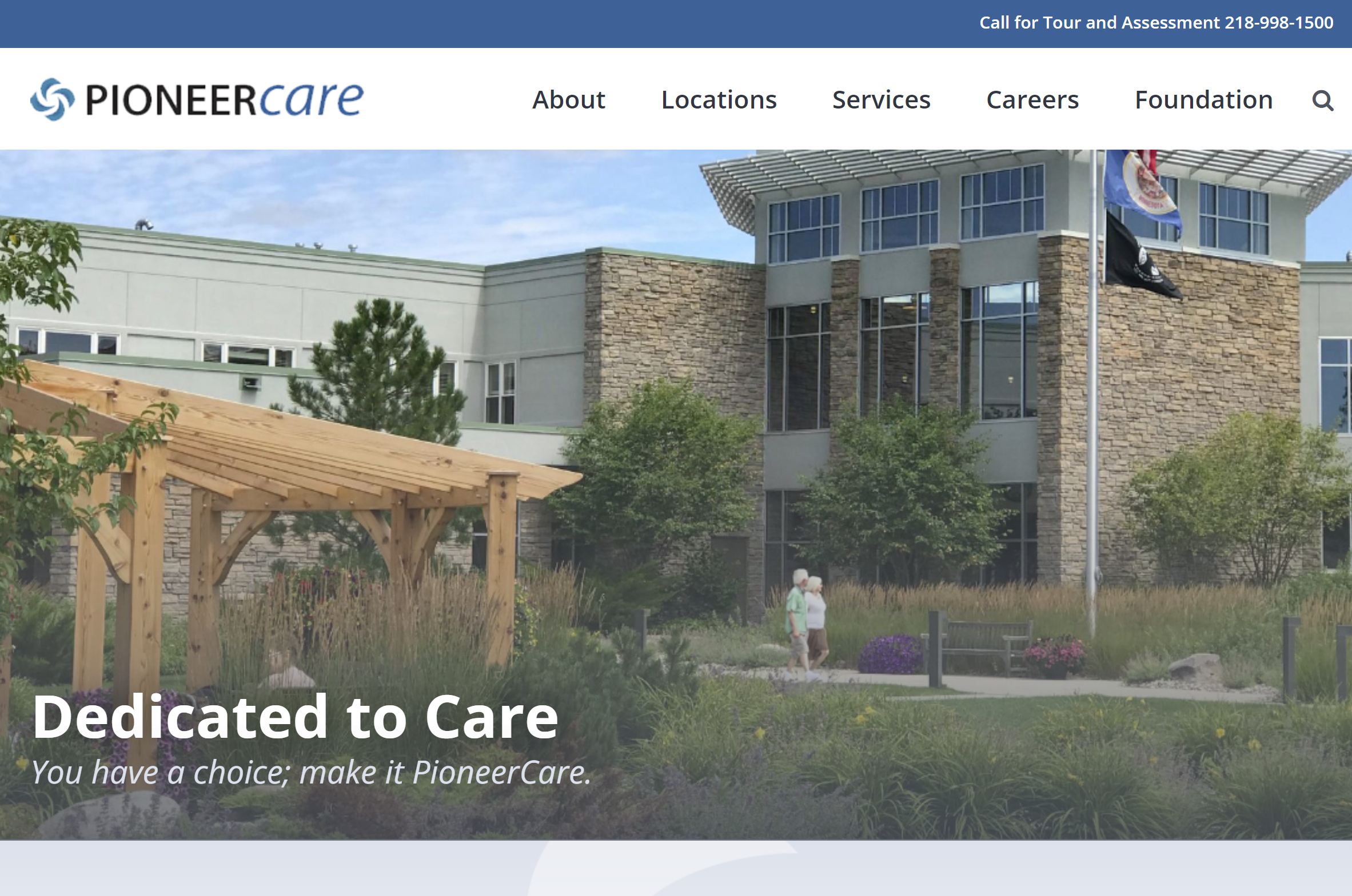 Front view of a modern building with stone and glass exterior, PioneerCare logo, and navigation menu. Showcasing the Portfolio of Pioneer Care Services, a wooden pergola and landscaped garden frame the entrance as two flags fly nearby.